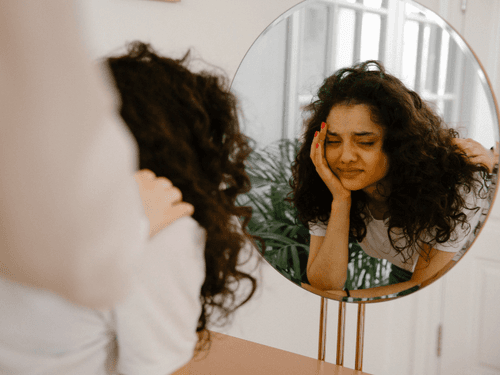 Mulher se olha no espelho com expressão de preocupação, apoiando o rosto, com uma mão visível atrás dela. Mulher se olha no espelho com expressão de preocupação, apoiando o rosto, com uma mão visível atrás dela.
